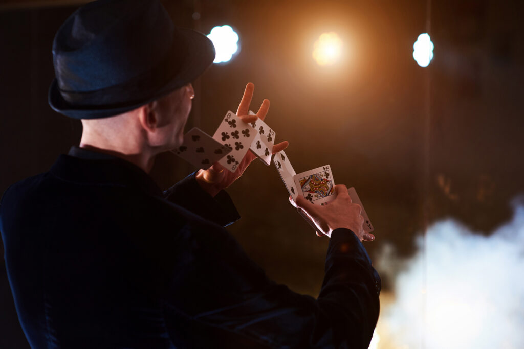 Magician showing trick with playing cards. Magic or dexterity, circus, gambling. Prestidigitator in dark room with fog Magician showing trick with playing cards. Magic or dexterity, circus, gambling. Prestidigitator in dark room with fog.